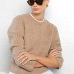 Woman wearing a beige sweater and sunglasses against a white brick wall.