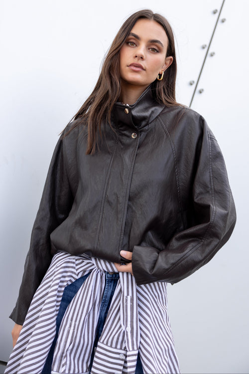 A woman wearing a black leather jacket with a high collar and front snap buttons, paired with a blue and white striped shirt tied at the waist, set against a light gray wall.