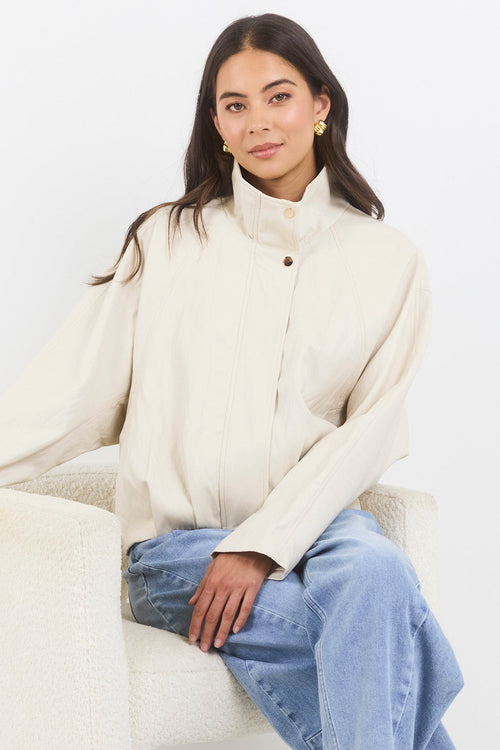 Woman wearing a cream coat and blue jeans sitting on a white couch.