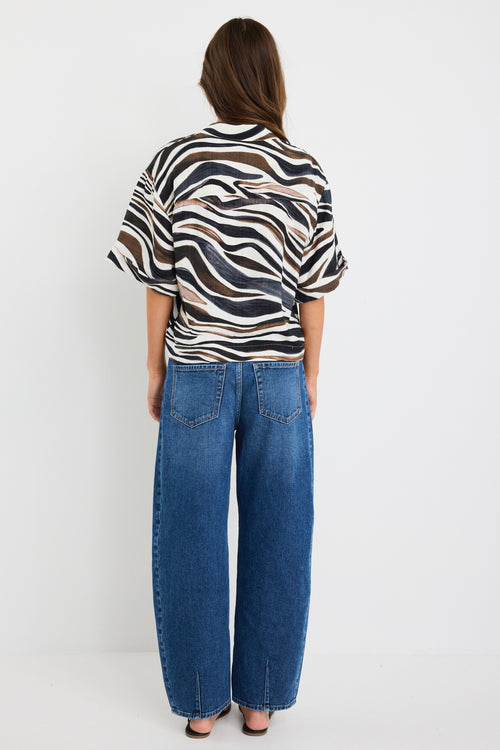 Woman wearing a relaxed-fit short sleeve zebra print shirt in chocolate, black, and white tones, styled with high-rise blue jeans.