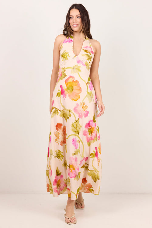 A woman modeling a sleeveless, halter-neck maxi dress with a vibrant pink, orange, and green floral print on a light beige background, set against a plain off-white studio backdrop.