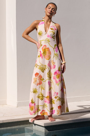 A woman models a sleeveless halter-neck maxi dress with a pink, green, and yellow floral watercolor print, standing on the edge of a swimming pool against a white stucco wall.