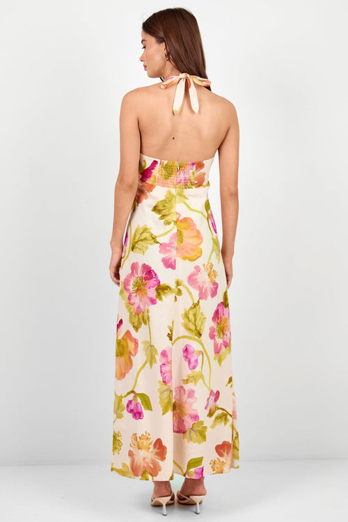 A woman is shown from the back wearing a halter-neck maxi dress with a vibrant floral print. The dress has a cream base with pink, orange, and green flowers.