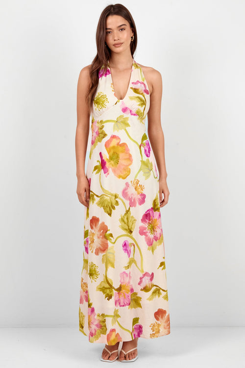 A woman modeling a sleeveless halter-neck maxi dress with a vibrant watercolor floral print on a cream background, standing against a plain light gray studio backdrop.