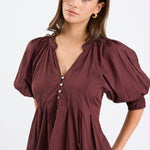 Woman wearing a chocolate brown pleated short-sleeve top with a button-down front, paired with blue jeans.