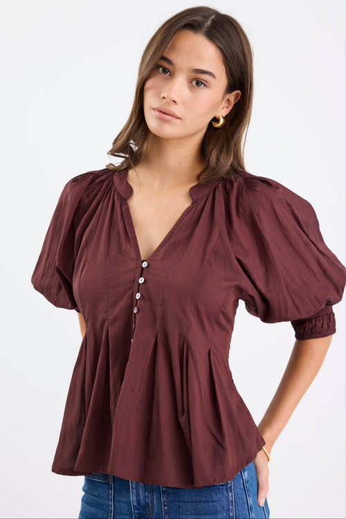 Woman wearing a chocolate brown pleated short-sleeve top with a button-down front, paired with blue jeans.