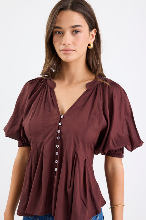 Woman wearing a chocolate brown pleated short-sleeve top with a button-down front, paired with blue jeans.