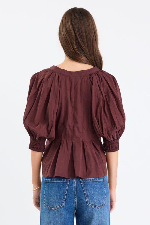 Woman wearing a chocolate brown pleated short-sleeve top with a button-down front, paired with blue jeans.