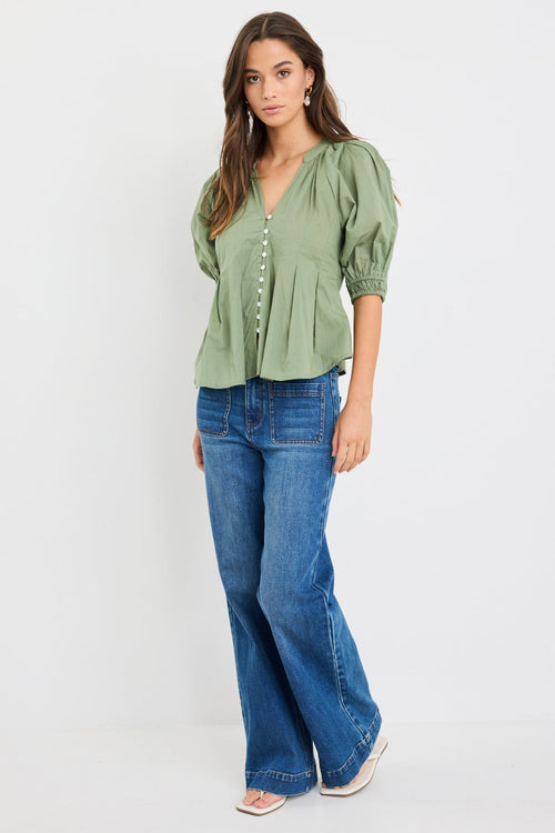 Woman wearing a moss green short sleeve button-front top with puff sleeves, pleated detail, and flared hem, styled with high-waist blue jeans.