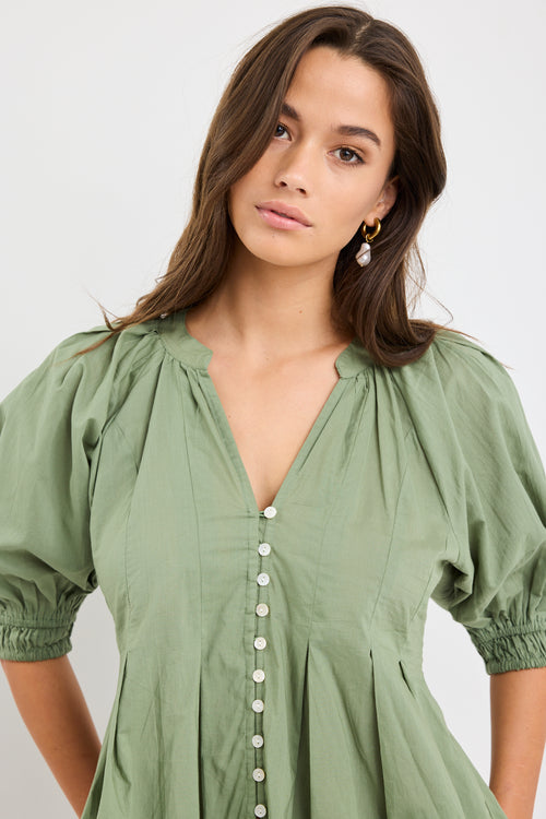 Woman wearing a moss green short sleeve button-front top with puff sleeves, pleated detail, and flared hem, styled with high-waist blue jeans.