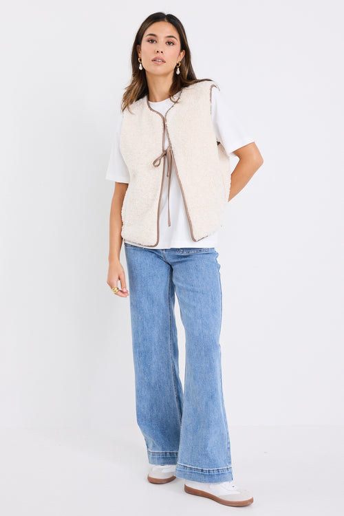 Woman wearing a fuzzy ecru sherpa gilet with contrast trim and front tie, styled over a white tee and light denim jeans.