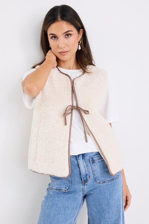 Woman wearing a fuzzy ecru sherpa gilet with contrast trim and front tie, styled over a white tee and light denim jeans.