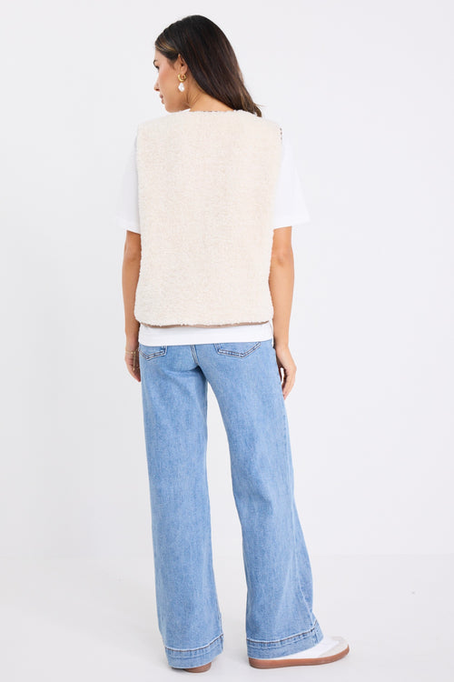 Woman wearing a fuzzy ecru sherpa gilet with contrast trim and front tie, styled over a white tee and light denim jeans.