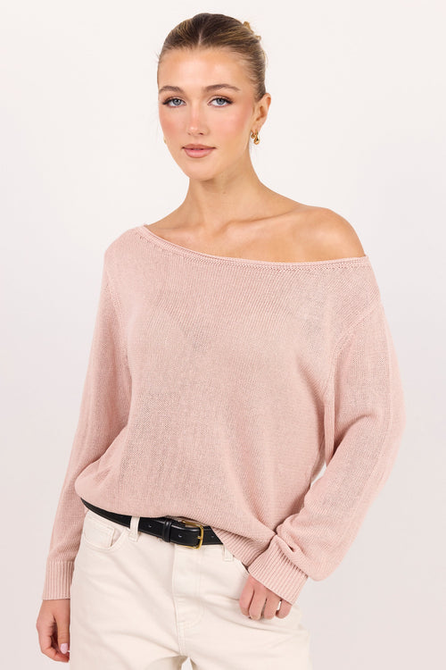 A woman modeling a light pink off-the-shoulder knit sweater with long sleeves, paired with white pants and a black belt, against a plain white studio background.