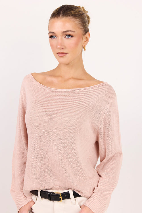 A woman wearing a pale pink knit sweater with a wide boat neckline, paired with white pants and a black belt, photographed against a plain light background.