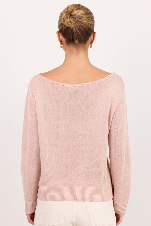 A woman viewed from the back wearing a pale pink knit sweater with a wide boat neckline and cream-colored trousers, set against a plain white studio background.