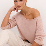 A woman wearing a dusty pink off-the-shoulder knit sweater, cream-colored pants, and black flat sandals, posing against a clean white studio background.