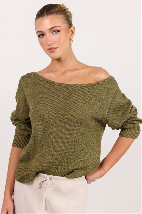A woman wearing an olive green off-the-shoulder knit sweater and cream drawstring pants, photographed against a clean off-white studio background.