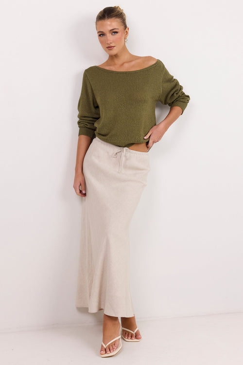 A woman modeling an olive green off-the-shoulder knit top and a cream-colored maxi skirt, standing against a plain white studio background.