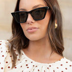 A woman wearing a cream puff-sleeve blouse with brown polka dots, styled with black sunglasses and blue jeans, sitting on outdoor stone steps.