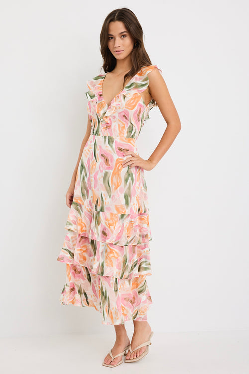 Woman wearing a sleeveless V-neck midi dress in a soft pink, peach, and green abstract print with a tiered skirt and tie detail at the back, styled with cream heeled sandals.