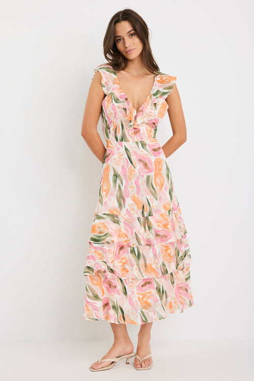 Woman wearing a sleeveless V-neck midi dress in a soft pink, peach, and green abstract print with a tiered skirt and tie detail at the back, styled with cream heeled sandals.