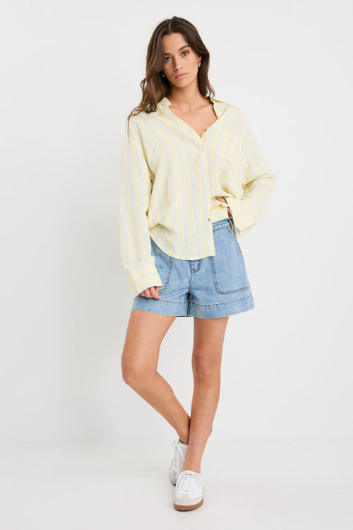 Woman wearing an oversized yellow and blue striped button-front shirt with chest pocket, styled with mid-blue denim shorts.