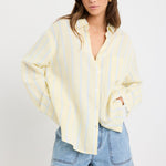 Woman wearing an oversized yellow and blue striped button-front shirt with chest pocket, styled with mid-blue denim shorts.