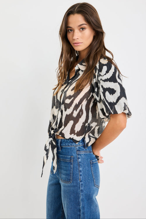 Woman wearing a black and cream zebra print short sleeve shirt with puff sleeves, button front, and tie waist detail, styled with blue jeans.
