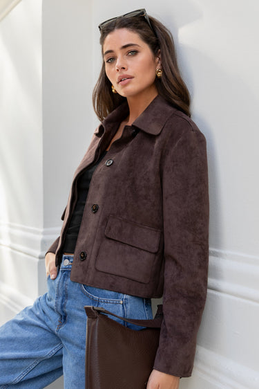Woman wearing a brown jacket and blue jeans against a white wall