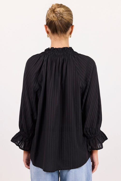 A woman viewed from the back wearing a black long-sleeve blouse with vertical pinstripes, a gathered smocked neckline, and ruffled cuffs, paired with light blue jeans against a plain white studio background.