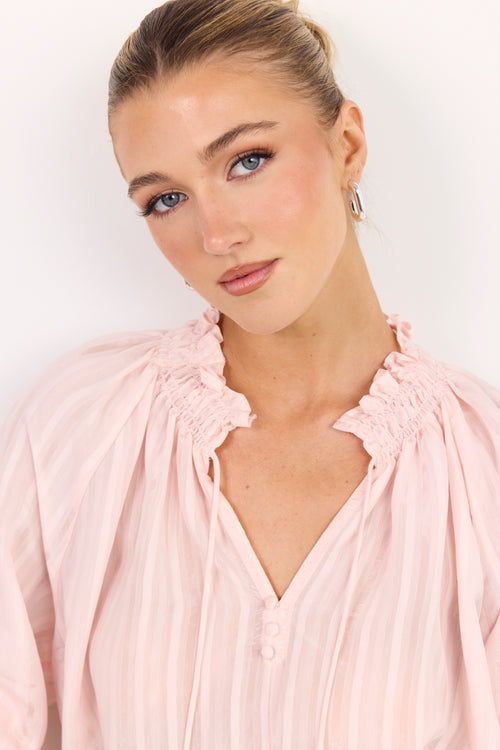 A close-up shot of a woman wearing a light pink blouse with ruffled details on the shoulders and a smocked neckline, set against a plain white background.