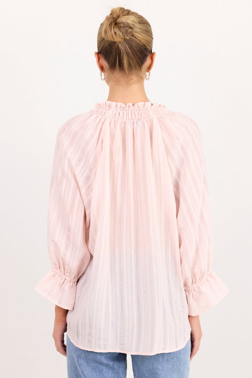A woman viewed from behind wearing a light pink pleated blouse with ruffled cuffs and a smocked neckline, paired with light blue jeans, against a plain white background.