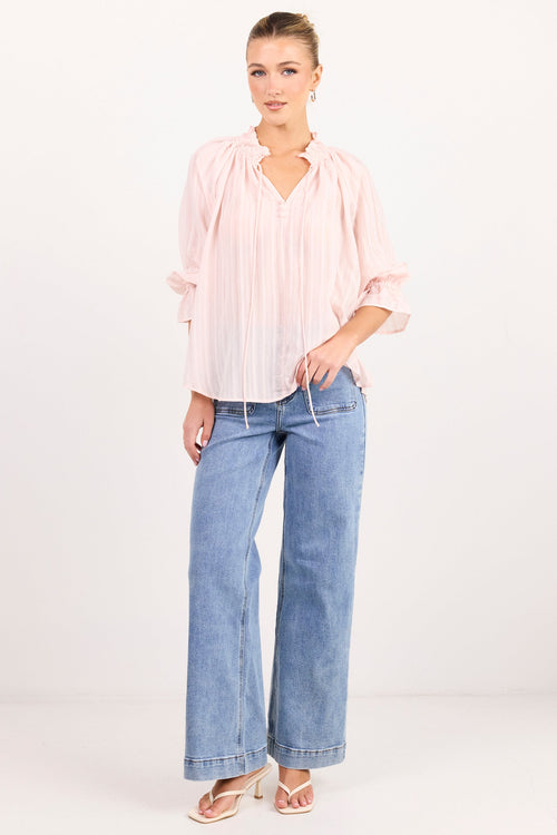 A woman modeling a light pink pleated blouse with three-quarter sleeves and light-wash blue wide-leg jeans, standing against a plain white studio background.