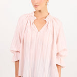 A woman modeling a light pink pleated blouse with a ruffled collar and three-quarter sleeves, paired with light blue jeans, against a plain white studio background.