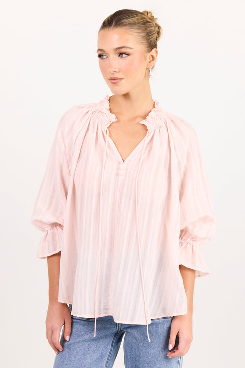 A woman modeling a light pink pleated blouse with a ruffled collar and three-quarter sleeves, paired with light blue jeans, against a plain white studio background.