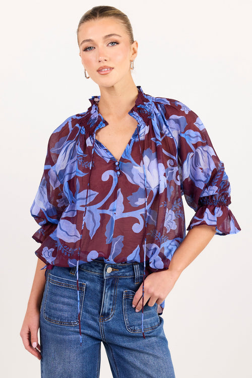 A woman modeling a sheer, ruffled blouse with a purple and blue floral pattern over a dark burgundy base, paired with blue jeans, against a plain white studio background.