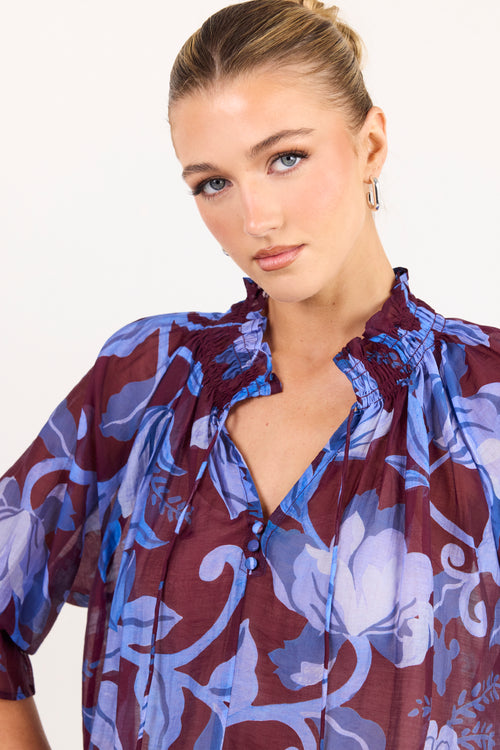 A woman wearing a sheer floral blouse with a deep V-neckline, featuring a pattern of blue and purple flowers over a dark burgundy background.