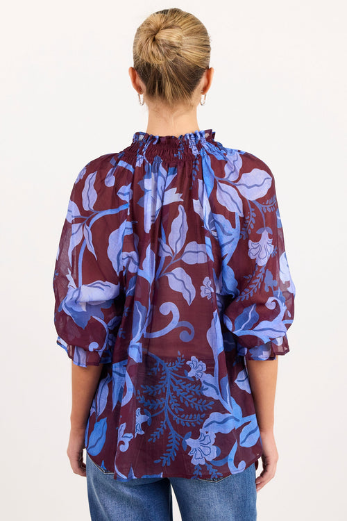 A woman viewed from behind wearing a sheer, long-sleeve blouse with a blue and purple floral pattern over a burgundy base, paired with blue jeans, against a plain white background.