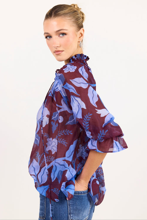 A woman wearing a sheer floral blouse with a deep purple base and blue floral print, paired with blue jeans, photographed against a plain white studio background.