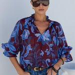 A woman modeling a sheer blue and burgundy floral print blouse with ruffled sleeves, paired with blue jeans and sunglasses, standing against a white wall.