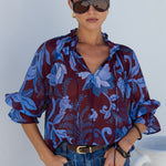 A woman modeling a sheer blue and burgundy floral print blouse with ruffled sleeves, paired with blue jeans and sunglasses, standing against a white wall.