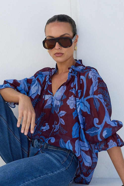 A woman wearing a blue and burgundy floral print blouse, blue jeans, and black sandals sits on a white platform against a white wall.