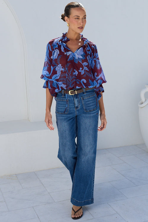 A woman stands outdoors against a white stucco wall wearing a blue and burgundy patterned blouse with ruffled sleeves, blue wide-leg jeans, and black sandals.