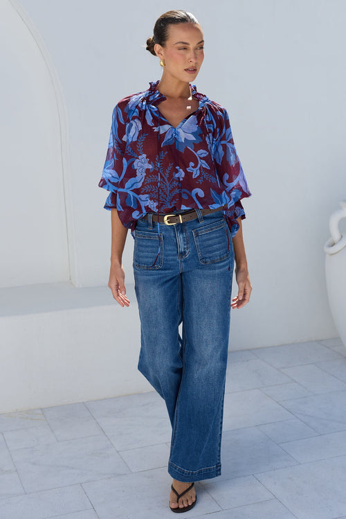 A woman stands outdoors against a white stucco wall wearing a blue and burgundy patterned blouse with ruffled sleeves, blue wide-leg jeans, and black sandals.