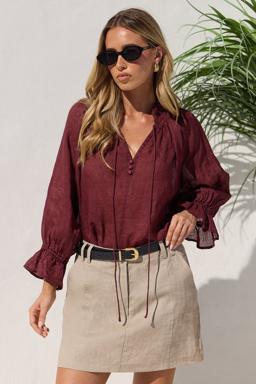 A woman modeling a burgundy long-sleeve blouse with ruffled cuffs and a tie-neck detail, paired with a beige linen mini skirt and a black belt, set against a white wall with a green plant in the background.