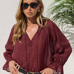 A woman wearing a burgundy long-sleeve blouse with a V-neck and gathered sleeves, paired with beige skirt and black sunglasses, standing against a light wall with a plant in the background.