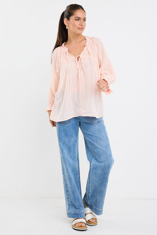 woman wearing a soft pink flowy cotton blouse with tie neckline and frill details, paired with blue jeans.