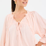 woman wearing a soft pink flowy cotton blouse with tie neckline and frill details, paired with blue jeans.