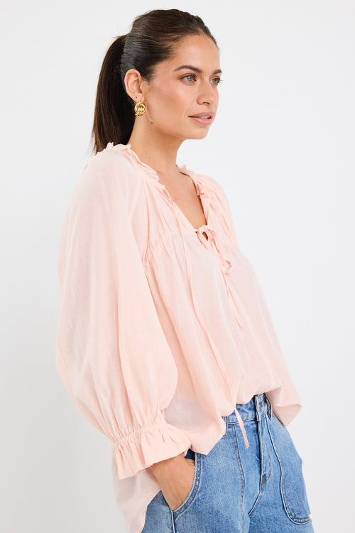 woman wearing a soft pink flowy cotton blouse with tie neckline and frill details, paired with blue jeans.